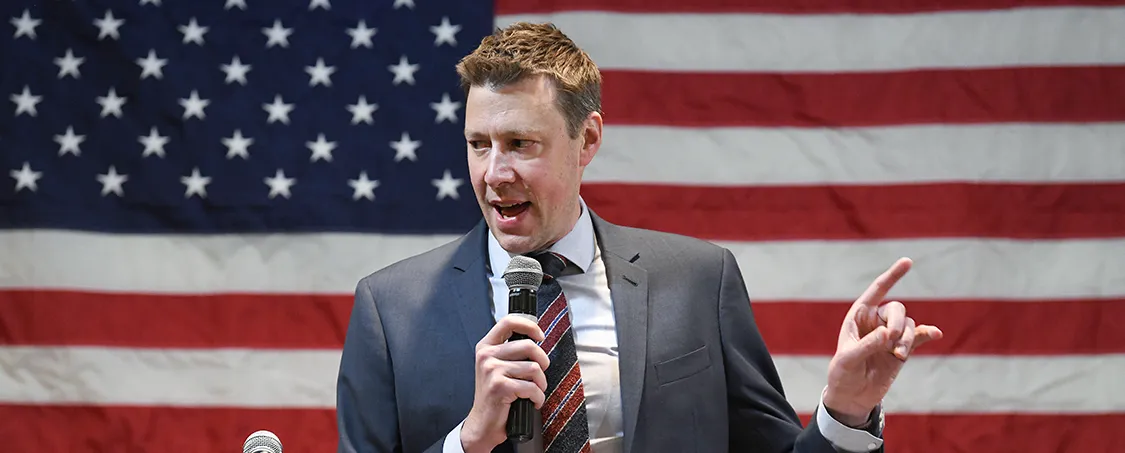 Person holding a microphone giving a speech on stage with a US lag behind him.